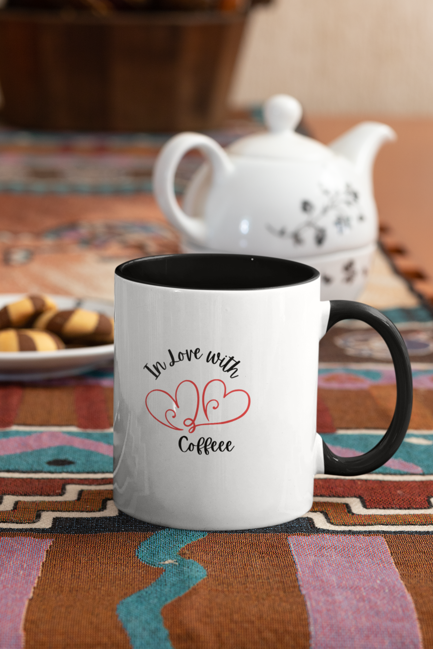 White mug with black interior and handle, featuring 'In Love with Coffee' text and heart design, on a colorful tablecloth with a teapot in the background.